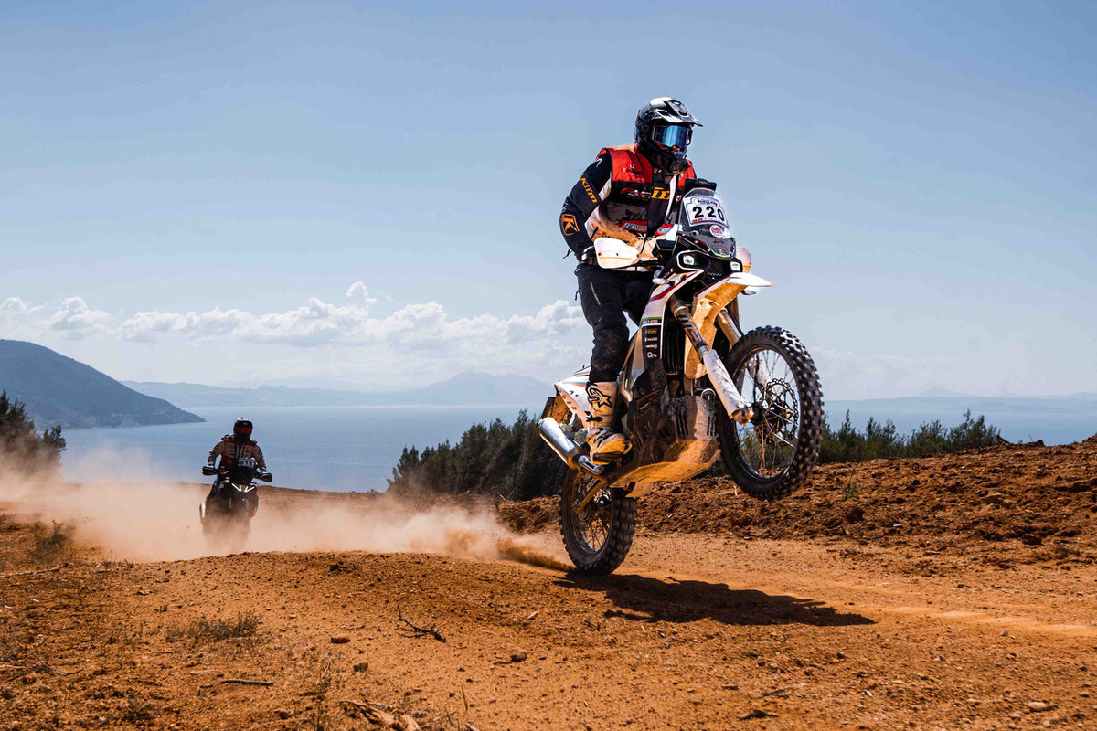 Pilota su moto rally impennata su uno sterrato di terra rossa in Sicilia, con uno specchio d'acqua sullo sfondo